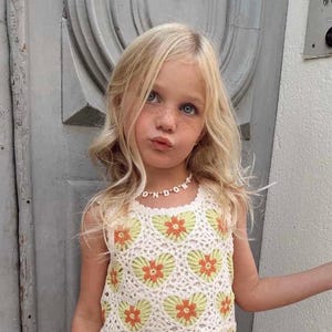 May include: A sleeveless crochet top featuring a repeating pattern of white, light green, and orange heart-shaped floral motifs. The top is paired with a beaded necklace that spells out the word LONDON in white letter beads on a string.