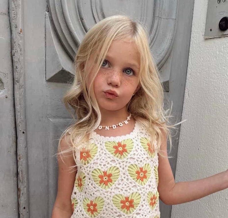 May include: A sleeveless crochet top featuring a repeating pattern of white, light green, and orange heart-shaped floral motifs. The top is paired with a beaded necklace that spells out the word LONDON in white letter beads on a string.