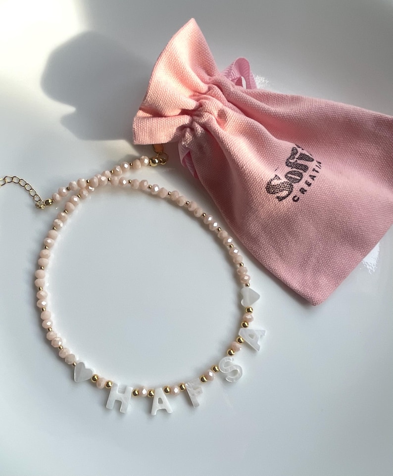 May include: A beaded necklace featuring light pink faceted beads, small gold-tone spacer beads, and white letter beads spelling HAFSA, accented with two white heart-shaped beads. The necklace is paired with a light pink fabric drawstring pouch.