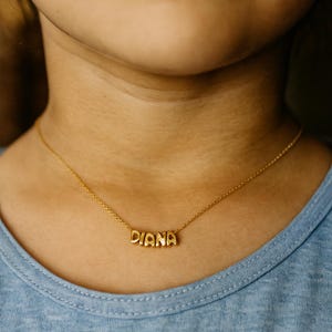 Könnte beinhalten: Eine goldfarbene Halskette mit dem Namen "DIANA" in Blockbuchstaben. Die Kette wird um den Hals einer Person getragen, im Hintergrund ist ein hellblaues Oberteil zu sehen. Ein zarter Schmuck.