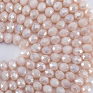 May include: A collection of pale pink, faceted glass beads with an iridescent finish. Each bead features a multi-sided, geometric cut that reflects light, creating a shimmering effect across the uniform, rounded shapes arranged in a dense cluster.