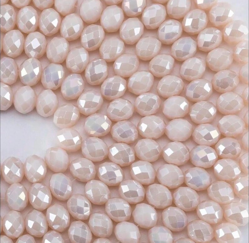 May include: A collection of pale pink, faceted glass beads with an iridescent finish. Each bead features a multi-sided, geometric cut that reflects light, creating a shimmering effect across the uniform, rounded shapes arranged in a dense cluster.