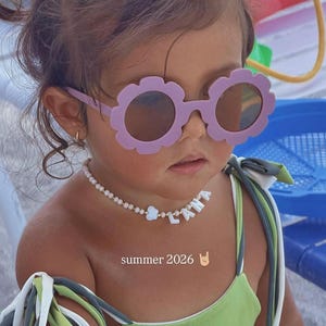 May include: Light purple flower-shaped sunglasses with dark lenses and a beaded necklace featuring white letter beads spelling LANA, a small cloud charm, and gold accents. The image includes the text summer 2026 with a hand gesture emoji.