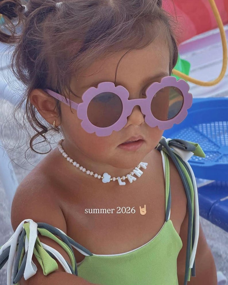 May include: Light purple flower-shaped sunglasses with dark lenses and a beaded necklace featuring white letter beads spelling LANA, a small cloud charm, and gold accents. The image includes the text summer 2026 with a hand gesture emoji.