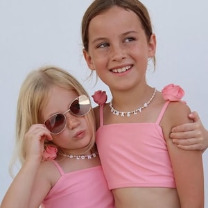 May include: Two pink crop tops featuring thin straps adorned with fabric roses at the shoulders. Each top is paired with a beaded necklace featuring white letter beads spelling out names, with one necklace reading BLAKELY and the other reading LONDON.