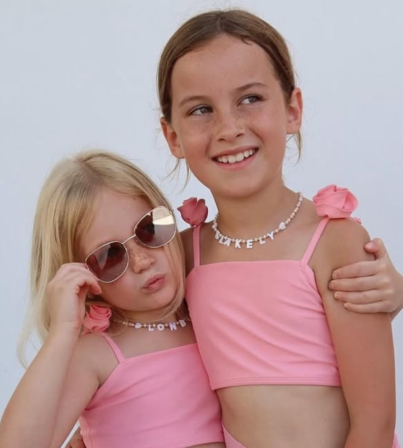 May include: Two pink crop tops featuring thin straps adorned with fabric roses at the shoulders. Each top is paired with a beaded necklace featuring white letter beads spelling out names, with one necklace reading BLAKELY and the other reading LONDON.