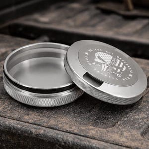 以下が含まれることがあります： 銀色の金属製の容器で、蓋が横に開いています。 蓋には、スカル、アメリカ国旗、および「WE ARE NOT DESCENDED FROM FEARFUL MEN 1776」のテキストがデザインされています。 容器にはテクスチャ加工されたエッジがあります。
