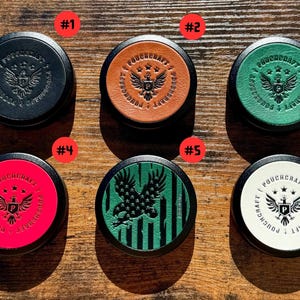 以下が含まれることがあります： 黒い縁が付いた、革製のトップが付いた6つの丸い容器。それぞれに、ワシ、星、および「POUCHCRAFT」のテキストなど、異なるデザインが施されています。色は黒、茶色、緑、赤、白などです。容器は木製の表面に表示されています。