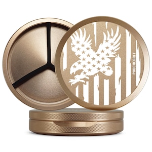 Op de afbeelding: Een goudkleurige, driedelige grinder met een patriottisch ontwerp. De bovenste deksel heeft een witte adelaar en sterren op een gestreepte achtergrond. De basis en het middengedeelte hebben een getextureerde afwerking. De tekst "POUCHCRAFT" is zichtbaar.