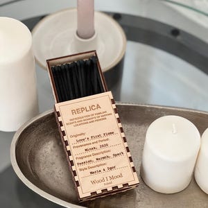 Op de afbeelding: Een houten doosje met lucifers met een zwart-wit label. Het label vermeldt "Replica" en bevat informatie over de geur, "Love's First Flame", de herkomst, "Minsk, 2020", en de geurbeschrijving, "Passie, Warmte, Vonk". Het label bevat ook de namen "Maria & Igor" en de merknaam "Wood I Mood, Polen".