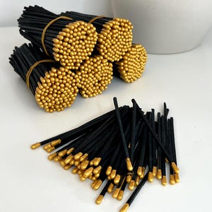 May include: Bundles of black matches with gold-colored tips, secured with rubber bands. Several bundles are stacked together, with a scattering of loose matches in the foreground. The matches are arranged on a white surface.