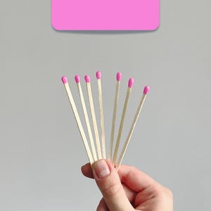 May include: A hand holding a fan of wooden matches with pink heads. The matches are set against a light grey background, with a pink rectangular shape at the top.