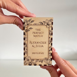 Op de afbeelding: Een rechthoekige houten doos met bloemengravures en de tekst "THE PERFECT MATCH" boven "ALEXANDER & JULIA" en de datum "24.05.2025". De doos wordt vastgehouden door twee handen.