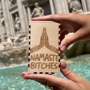 May include: A rectangular wooden box with the phrase "NAMASTE BITCHES" etched on the front. The design features hands in a prayer position. The box is held in front of a blurred background of a fountain and statues.