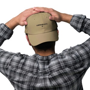 Può includere: Un berretto da baseball beige con la scritta "ADDERALL" stampata in nero. Il berretto è tenuto da una persona che indossa una camicia a quadri bianca e nera.