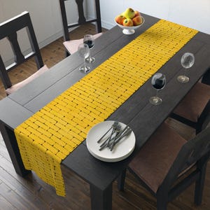 May include: A yellow table runner with a brick pattern is centered on a dark wooden dining table. The table is set with plates, silverware, and wine glasses. A bowl of fruit sits at the end of the table.