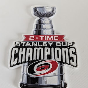 May include: A flat, die-cut emblem featuring the Stanley Cup trophy in silver and black. A red banner across the center reads 2-TIME STANLEY CUP CHAMPIONS in white text. The Carolina Hurricanes logo is centered over the base of the trophy.