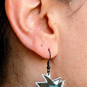 May include: A black earring hook holding a white, black, and blue hockey-themed charm. The charm features a stylized shark logo with a hockey stick and a blue and black color scheme. The earring is worn on a person's ear.