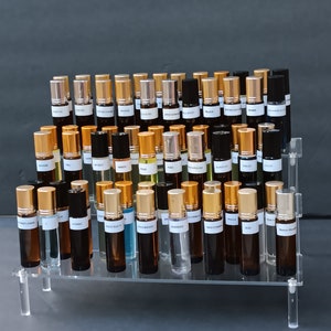 May include: A clear acrylic display stand holding numerous small perfume or essential oil bottles. The bottles are various shades of brown and clear glass, with gold or black caps. Each bottle has a white label with black text.