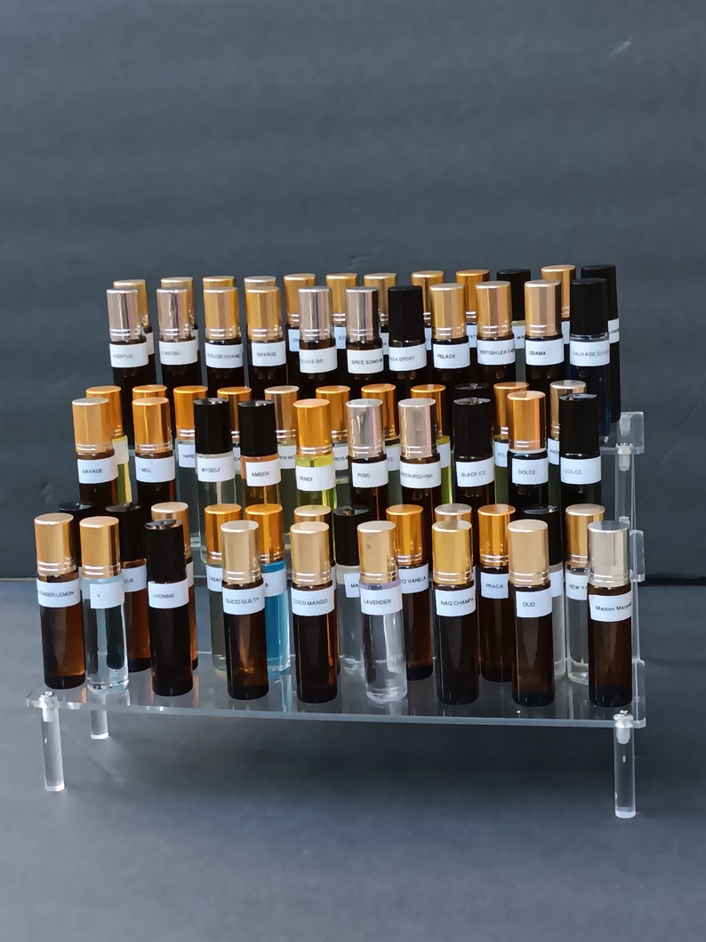 May include: A clear acrylic display stand holding numerous small perfume or essential oil bottles. The bottles are various shades of brown and clear glass, with gold or black caps. Each bottle has a white label with black text.