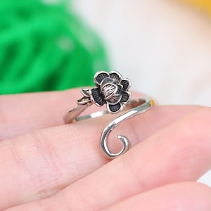 Op de afbeelding: Een zilverkleurige ring met een bloemmotief. De ring heeft een donkere, gedetailleerde bloem met bladeren en een gebogen band. De ring wordt getoond op de hand van een persoon, met een vage groene en gele achtergrond.