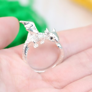 May include: A silver-colored dragon ring with textured details. The dragon has outstretched wings and a small, dark-colored eye. The ring is shown on a hand, with a blurred background.