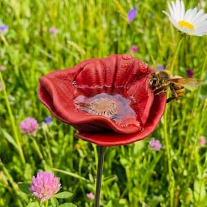 Peut inclure: Un objet sculpté en forme de fleur rouge foncé avec une dépression centrale contenant de l'eau et un centre beige texturé. Une abeille est posée sur le bord des pétales rouges, qui présentent des gouttelettes d'eau, fixée sur une tige fine.