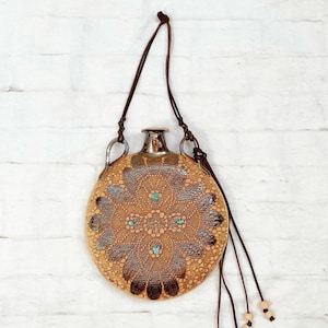 Puede incluir: Una cantimplora redonda y decorativa con exterior de cuero marrón y parte superior de metal plateado. La cantimplora presenta un diseño simétrico detallado en tonos marrón, beige y turquesa. Una correa de cuero marrón y borlas de cuentas cuelgan de la cantimplora.
