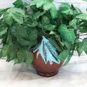 Op de afbeelding: Een kunstplantarrangement in een bruine keramische pot. De pot heeft een lichtblauw bladmotief. De plant heeft groene bladeren. De achtergrond is een witte bakstenen muur.