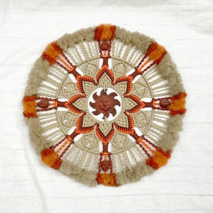 May include: Circular macrame wall hanging with a sun-like design. The center features a brown sun face, surrounded by a floral pattern in beige and orange. The outer edge has a fluffy, light brown texture with orange accents.