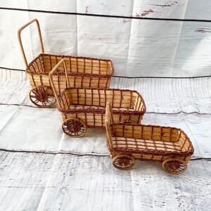May include: Three miniature woven wicker wagons with brown accents. Each wagon has a rectangular basket, a handle, and two wheels. The wagons are arranged in a descending size order, showcasing a rustic, handcrafted aesthetic.