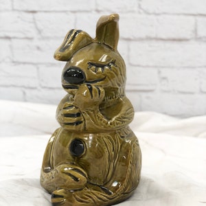 May include: A ceramic figurine of a seated rabbit with closed eyes, in a light olive green glaze. The rabbit has black accents on its nose, paws, and ears. The figurine is detailed with textured fur and a button on its chest.
