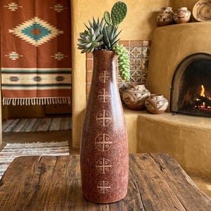 May include: A tall, reddish-brown ceramic vase with a series of white cross-like designs, holding succulents including a prickly pear cactus. The vase is on a rustic wooden table. The room has a Southwestern style, with a terracotta colour scheme.