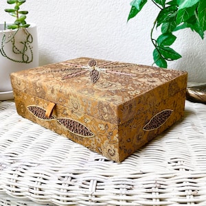 Könnte beinhalten: Eine rechteckige, goldfarbene Schmuckschatulle mit floralem Muster und einem Libellen-Design auf dem Deckel. Die Schatulle hat Perlenakzente und einen kleinen Verschlussriemen in Beige. Die Schatulle steht auf einer weißen Korboberfläche.