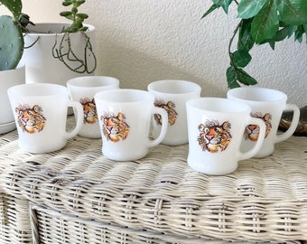 May include: A set of six white ceramic mugs with a tiger face design. The mugs have a curved handle and a slightly flared rim. The tiger design features orange, brown, and yellow colors. The mugs are arranged on a white wicker surface.