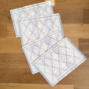 May include: Four rectangular placemats with a geometric diamond pattern in pastel colors of pink, blue, and white. Each placemat has a white border with a fringe edge. The placemats are arranged on a wooden surface.