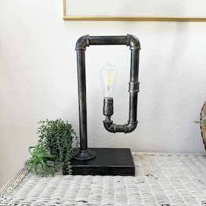 May include: Industrial-style table lamp with a black wooden base and a frame made of dark gray metal pipes. The lamp features a vintage-style Edison bulb. The lamp is designed to sit on a table or desk.