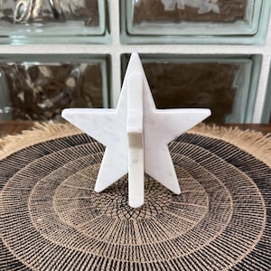 May include: A white marble star-shaped decorative object. The star is standing upright, with a vertical support piece. The object is set on a round, patterned mat with a fringed edge. The background includes glass block windows.