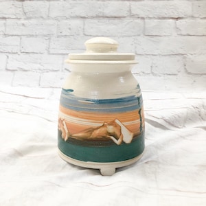 May include: A ceramic jar with a lid, featuring a landscape design. The jar has a cream-colored base with a painted scene of mountains, blue and orange stripes, and a green base. The jar is approximately 8 inches tall.