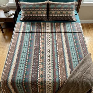 Może przedstawiać: Łóżko z turkusową pościelą, dwiema poszewkami w wzory i pasującą wzorzystą kołdrą. Kołdra i poszewki mają geometryczny wzór w odcieniach brązu, turkusu i beżu. Brązowy koc jest narzucony na łóżko.