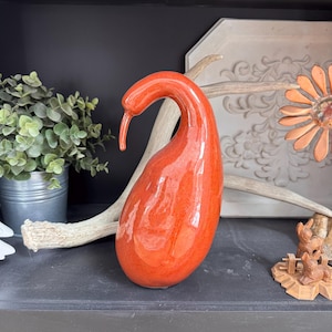 May include: A glossy, orange ceramic sculpture with a curved neck and beak-like tip. The abstract form sits on a dark surface, with a metal bucket of greenery and decorative elements in the background. The sculpture's color is a vibrant, reddish-orange.