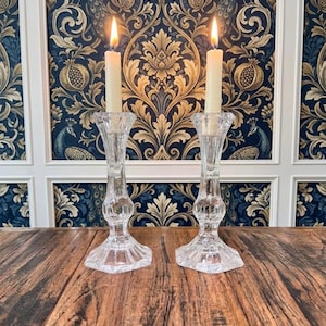May include: Two clear glass candle holders with faceted stems and wide, hexagonal bases. Each holder supports a single off-white taper candle with a lit flame. The glass features a reflective, cut-crystal design that catches the light.