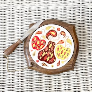 Puede incluir: Una tabla de quesos vintage con una baldosa de cerámica decorativa con frutas estilizadas en rojo, marrón y amarillo con detalles dorados. La tabla tiene un marco de madera e incluye un cuchillo de queso con mango de madera y cadena de metal.