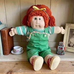 May include: A soft-sculpture doll with bright orange yarn hair tied with a yellow ribbon, wearing a green and white striped shirt under green overalls. The doll has green eyes, a round face, and wears white sneakers with red stripes on the sides.