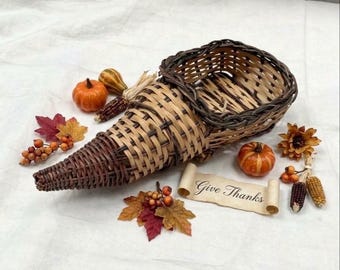 May include: A woven wicker cornucopia basket featuring dark and light brown reeds. It is surrounded by decorative autumn elements including small orange pumpkins, colorful fabric leaves, clusters of berries, corn cobs, and a scroll reading Give Thanks.