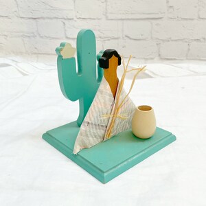 Pode incluir: Uma peça decorativa de madeira turquesa com um cacto, uma figura nativa americana e um pequeno vaso. A figura é coberta por um tecido estampado e adornada com palha. A base é quadrada e pintada de turquesa.