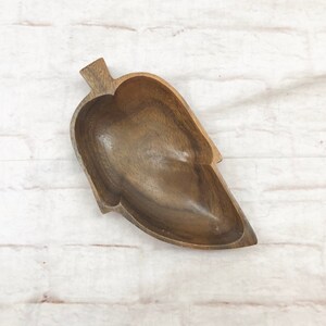 Pode incluir: Uma tigela de madeira em forma de folha com acabamento natural de grão de madeira. A tigela tem um interior profundo e uma alça em forma de haste. A tigela tem aproximadamente 25 cm de comprimento e 15 cm de largura, perfeita para servir lanches ou exibir itens decorativos.