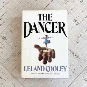 May include: A book titled "The Dancer" by Leland Cooley. The cover shows a ballerina in a blue dress balanced on a hand-shaped sculpture. The title is in bold black letters.