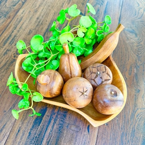 May include: A wooden serving tray holds carved wooden fruit and decorative greenery. The tray contains a pear, a banana, and several round fruits with star and geometric designs. The wooden fruit is a warm brown color.