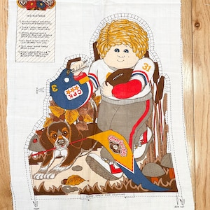 Könnte beinhalten: Weißes Stoffpaneel mit einem Cabbage Patch Kids-Design. Die Illustration zeigt ein Kind in Football-Uniform mit einem Football und einem Hund. Enthält Anweisungen und den Text "Cabbage Patch Kids".